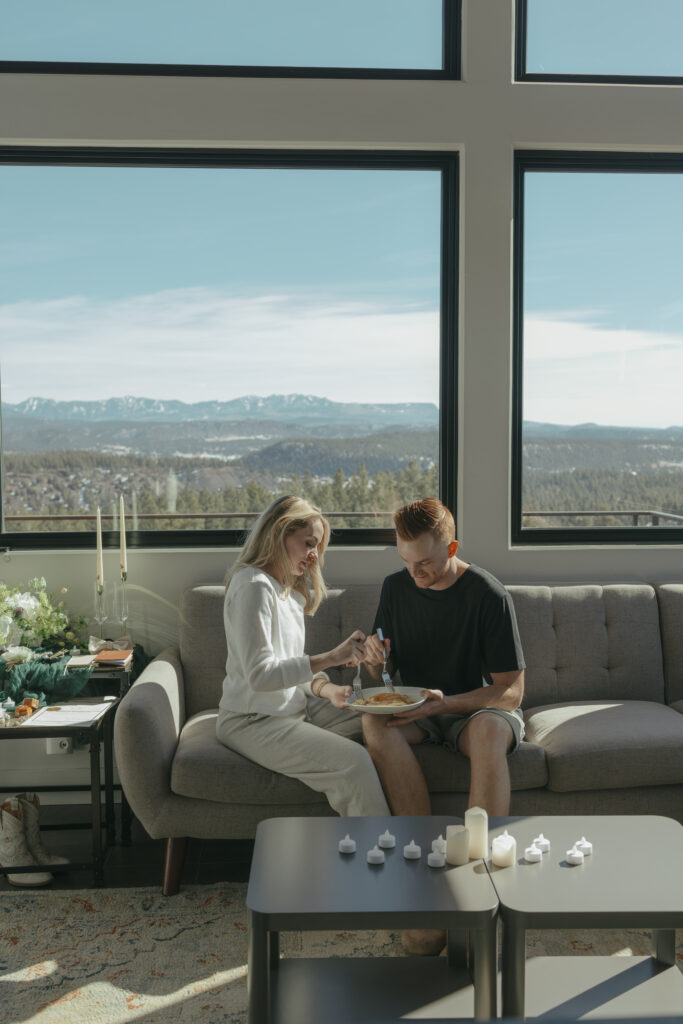 Bride and Groom snuggled up on the couch together, sharing pancakes, with the mountain backdrop on their elopement day