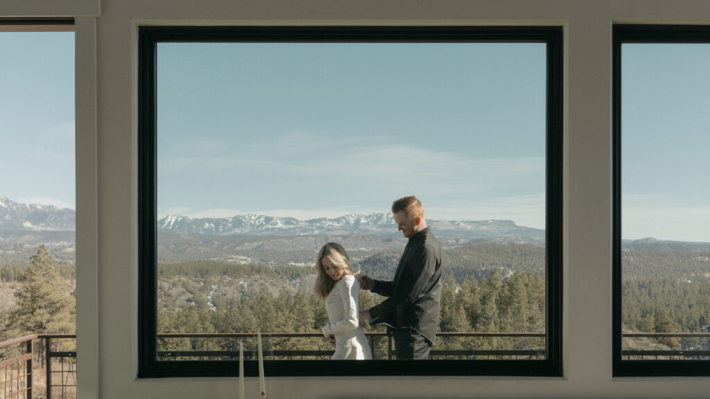 Groom helping bride zip up her dress as they stand on the balcony of their Airbnb overlooking the colorado mountains 