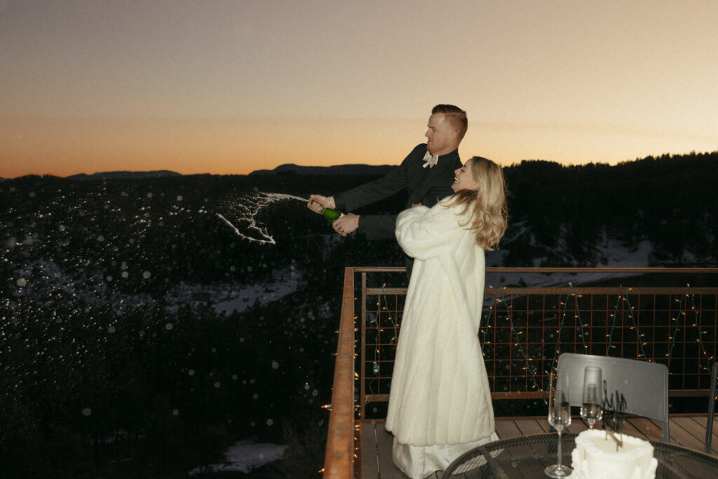 Bride and groom standing on balcony popping bottle of champagne at sunset 