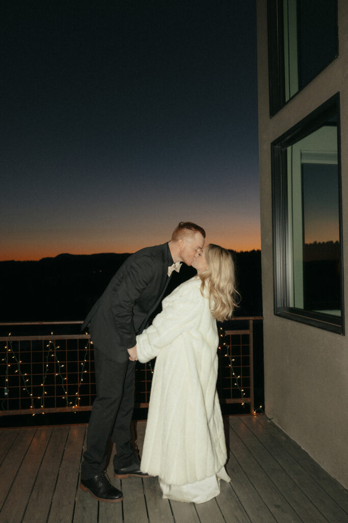 Bride and groom kissing on balcony overlooking mountains at sunset