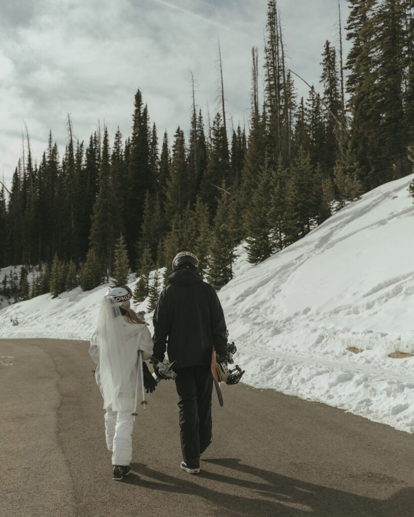 Bride and groom in their ski gear, with veil attached to helmet, holding their snowboard and skis while walking