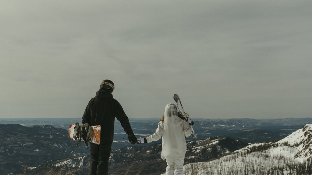 bride and groom overlooking the mountains while holding their snowboard and skis at the top of Wolf Creek Ski Area in Pagosa Springs Colorado