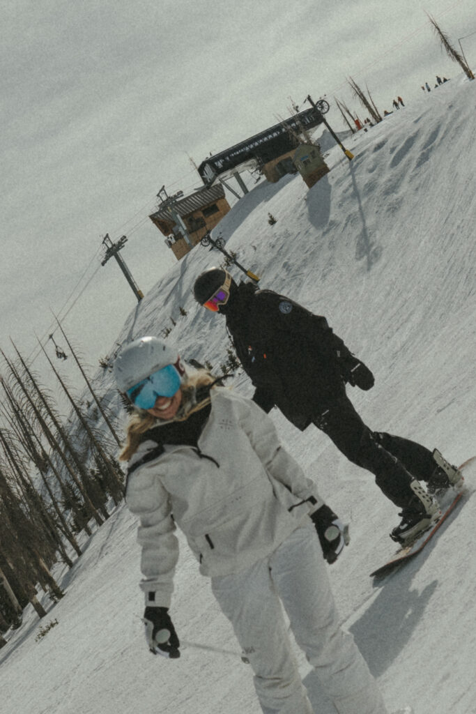 Bride smiling while skiing down the mountain and groom chasing her on his snowboard