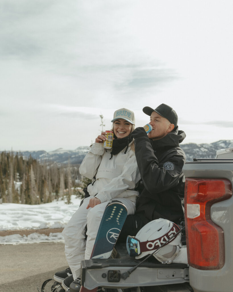 Bride sitting in grooms lap on tailgate drinking a beer 