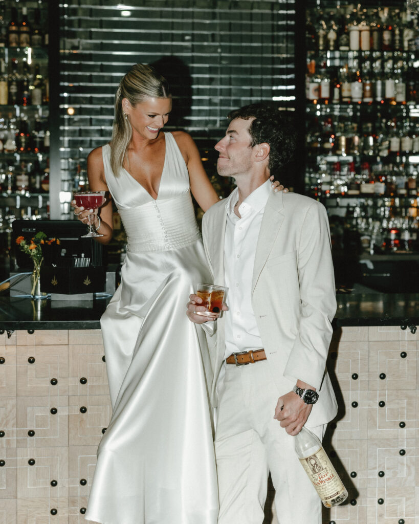 bride sitting on top of the bar while holding her cocktail with groom holding a bottle of pappy Van winkle