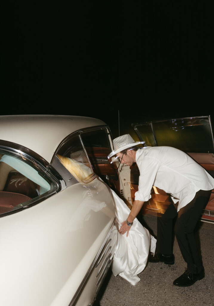 Groom helping bride into vintage car during wedding send-off at Trinity View Farm