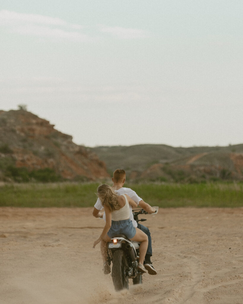 Couple riding a dirt bike through sandy landscape during a moody Texas engagement session