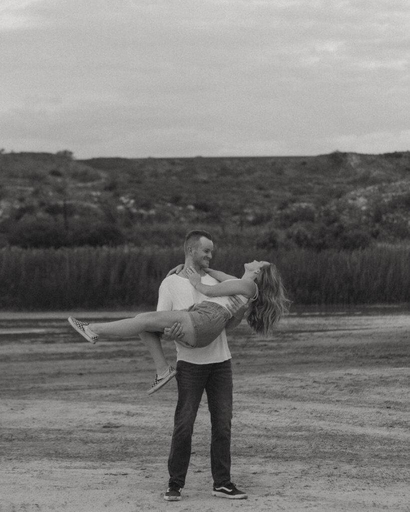 Black and white engagement photo of groom lifting bride in open Texas landscape